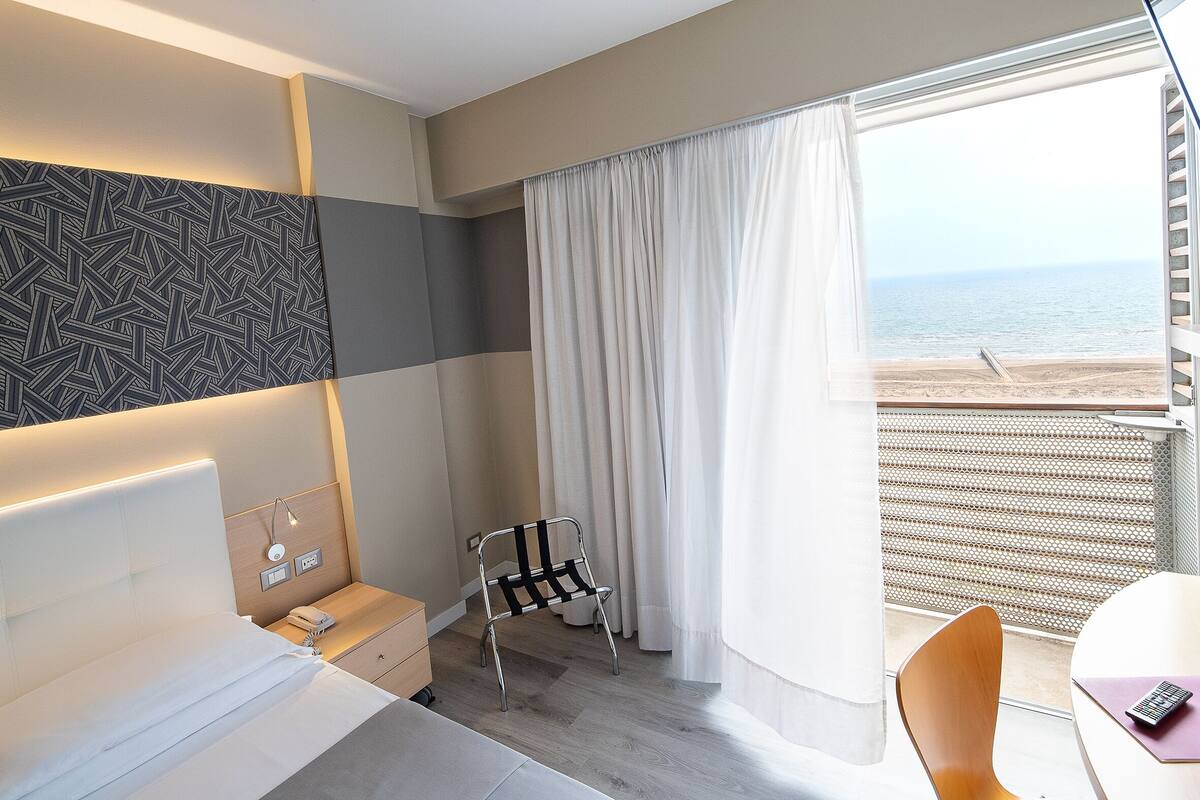 Een hotelkamer met een bed, nachtkastje, stoel en uitzicht op het strand door een groot raam.