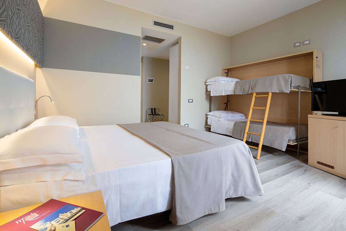Een hotelkamer met een enkel bed, een stapelbed, een bureau met een boek, een televisie en een kledingkast.