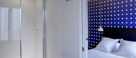 Un baño moderno con cabina de ducha de vidrio y una cama con ropa de cama blanca y un cabecero azul estampado.