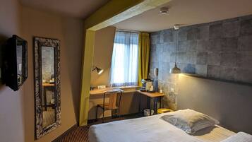 A hotel room with a large bed, a desk with a chair, a mirror, and a window with curtains.