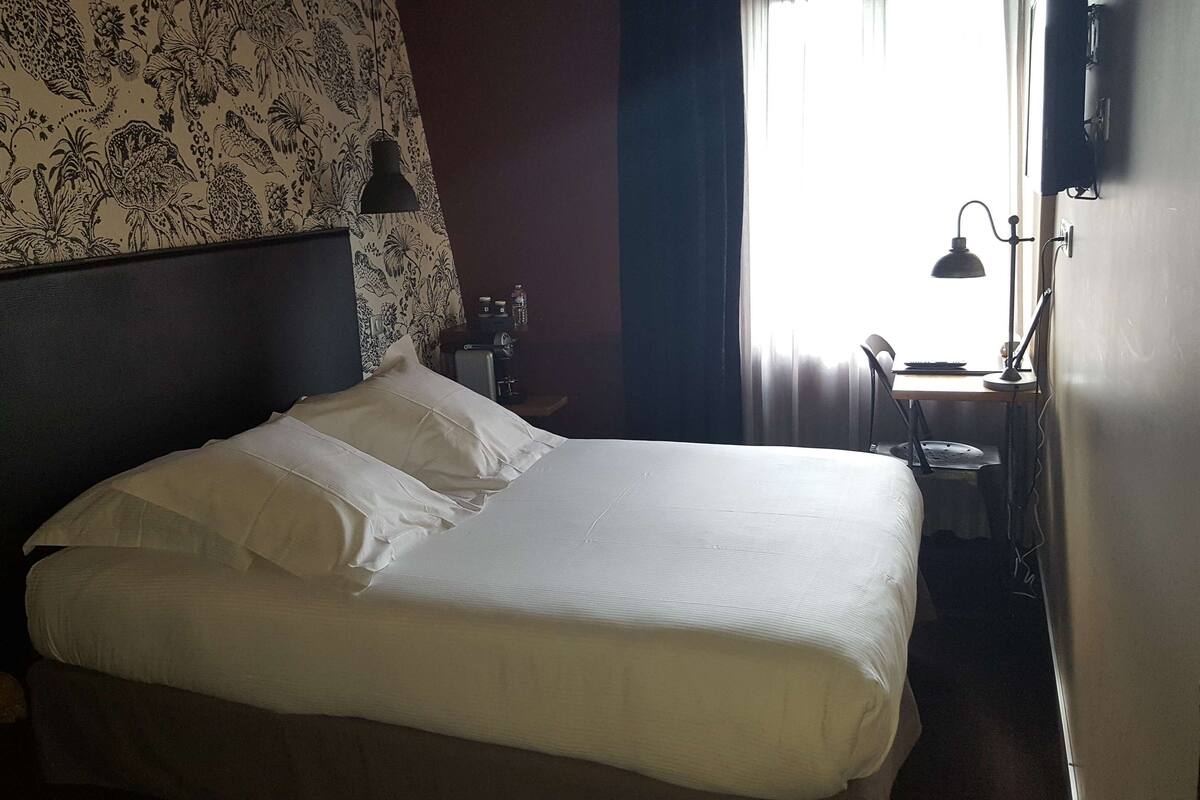 A neatly made bed with white linens in a room with floral wallpaper, a dark headboard, and a desk with a lamp and a bottle.