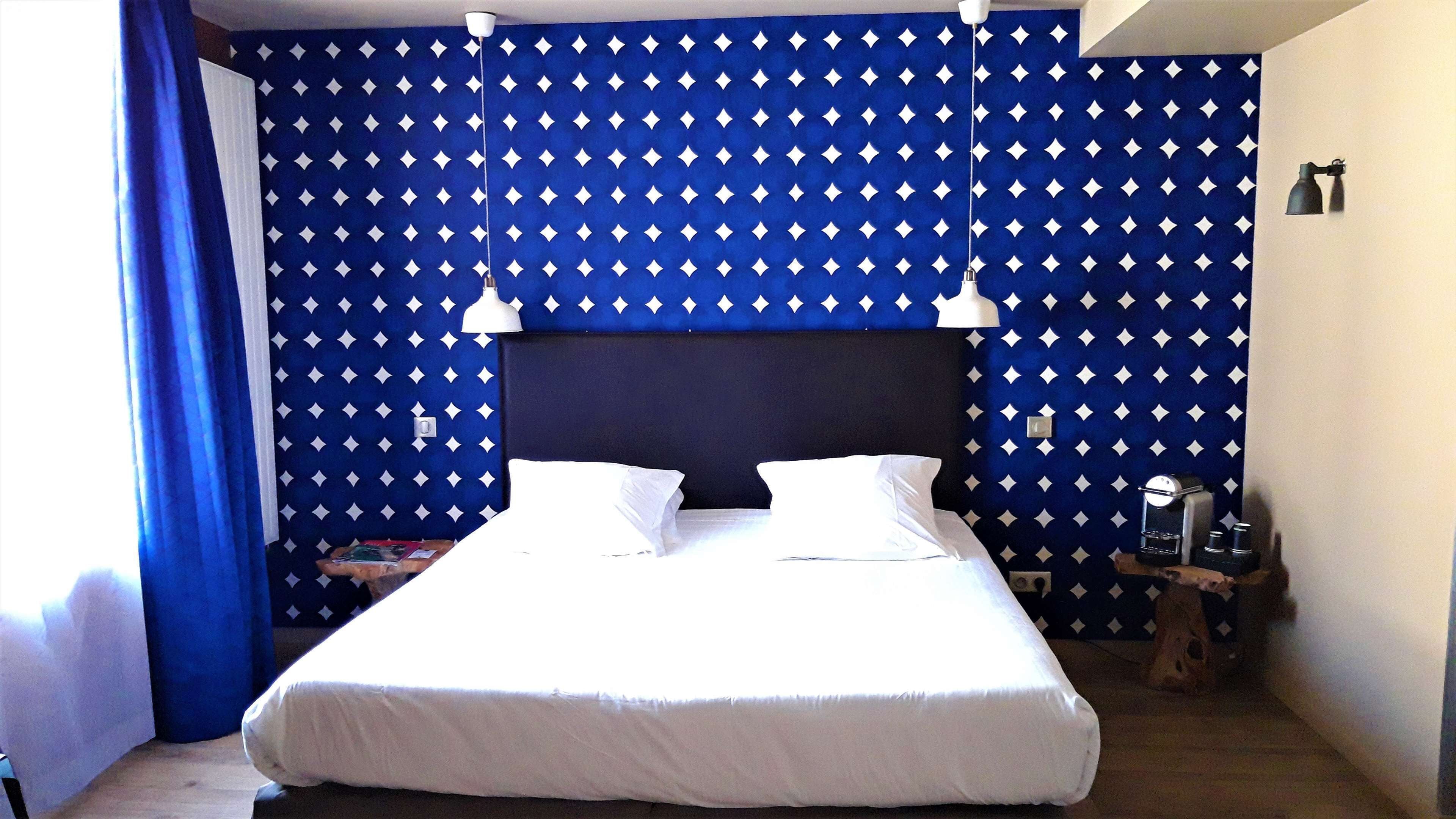 Un lit bien fait, avec du linge de lit blanc, dans une pièce dont un mur est décoré d’un motif bleu et qui est agrémenté d’une tête de lit sombre.