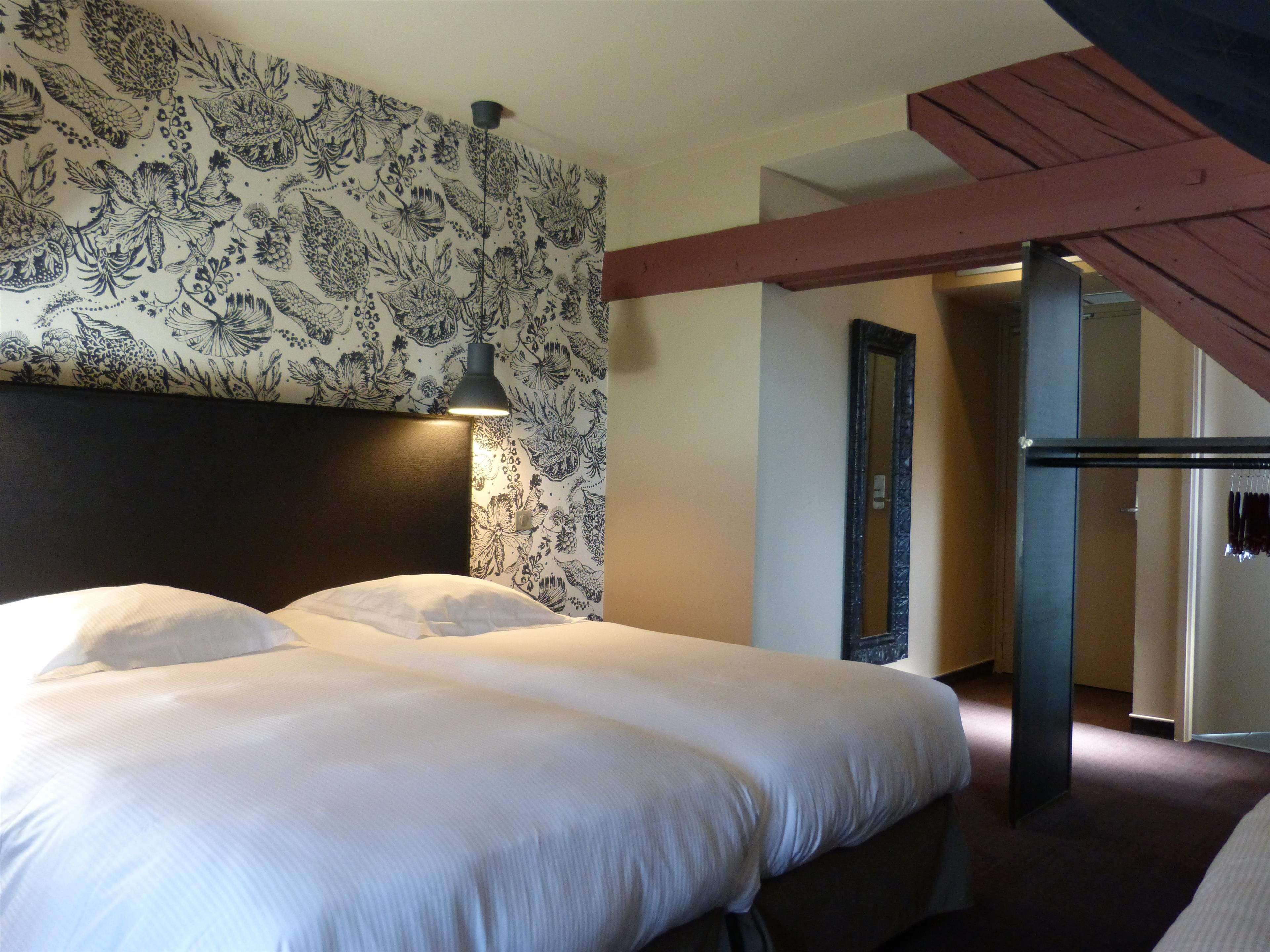 Une chambre d’hôtel avec un grand lit, une tête de lit recouverte d’un papier peint à motifs floraux, une lampe suspendue et une poutre en bois au plafond.