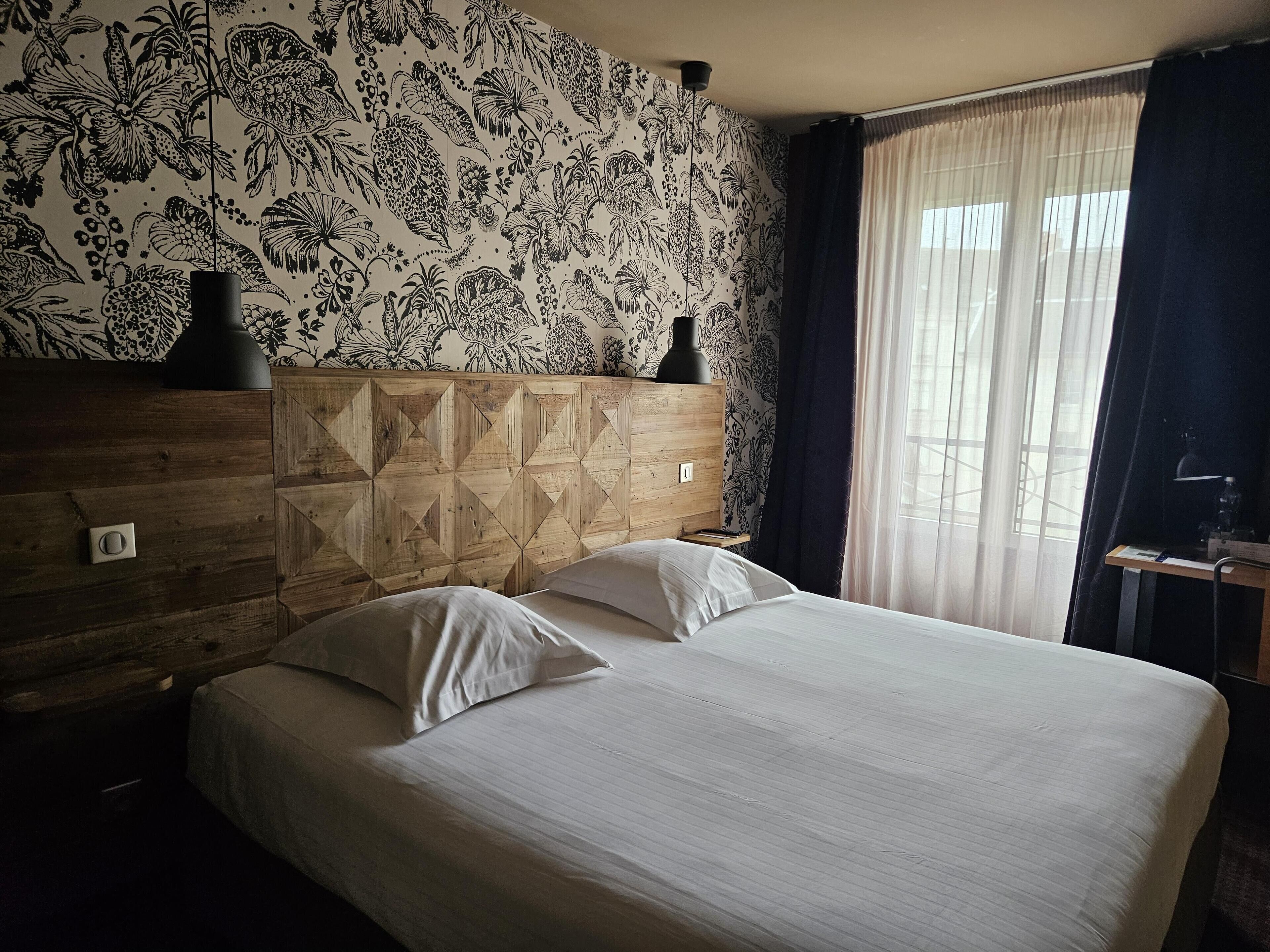 A bed with white bedding, a wooden headboard with geometric patterns, a nightstand, a lamp, and a window with sheer curtains.