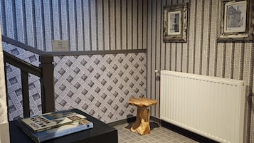 A room with a patterned wallpaper, a radiator, a wooden stool, and a table with books.