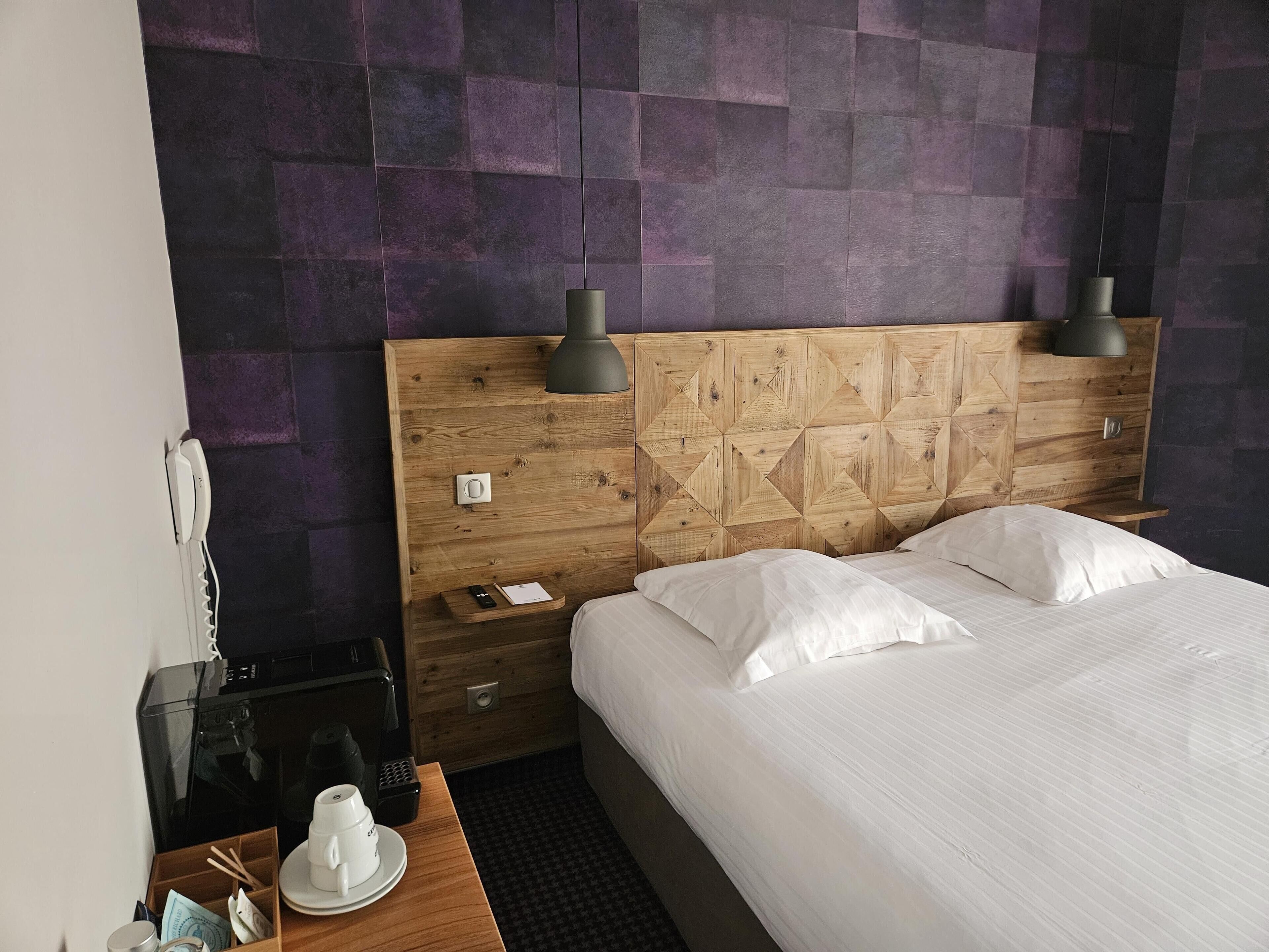 Une chambre d’hôtel avec une tête de lit en bois, deux lampes de chevet, un lit avec du linge de lit blanc, une table de chevet avec un téléphone, une machine à café et une tasse.