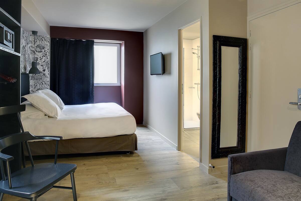 A bedroom with a bed, a chair, a TV, and a door leading to a bathroom.
