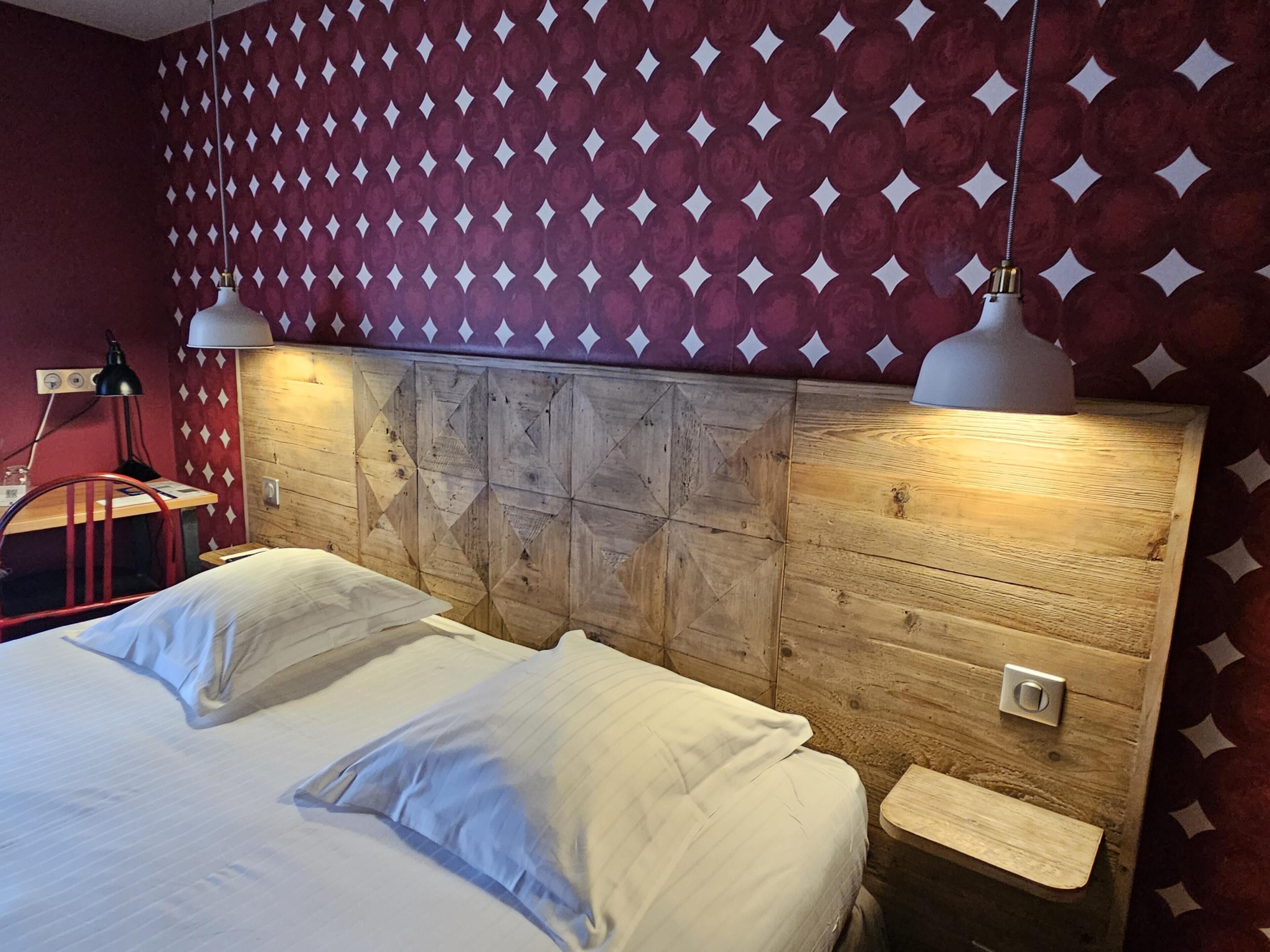 Une chambre avec une tête de lit en bois, un lit recouvert de draps blancs, une chaise rouge et un mur orné d’un motif.