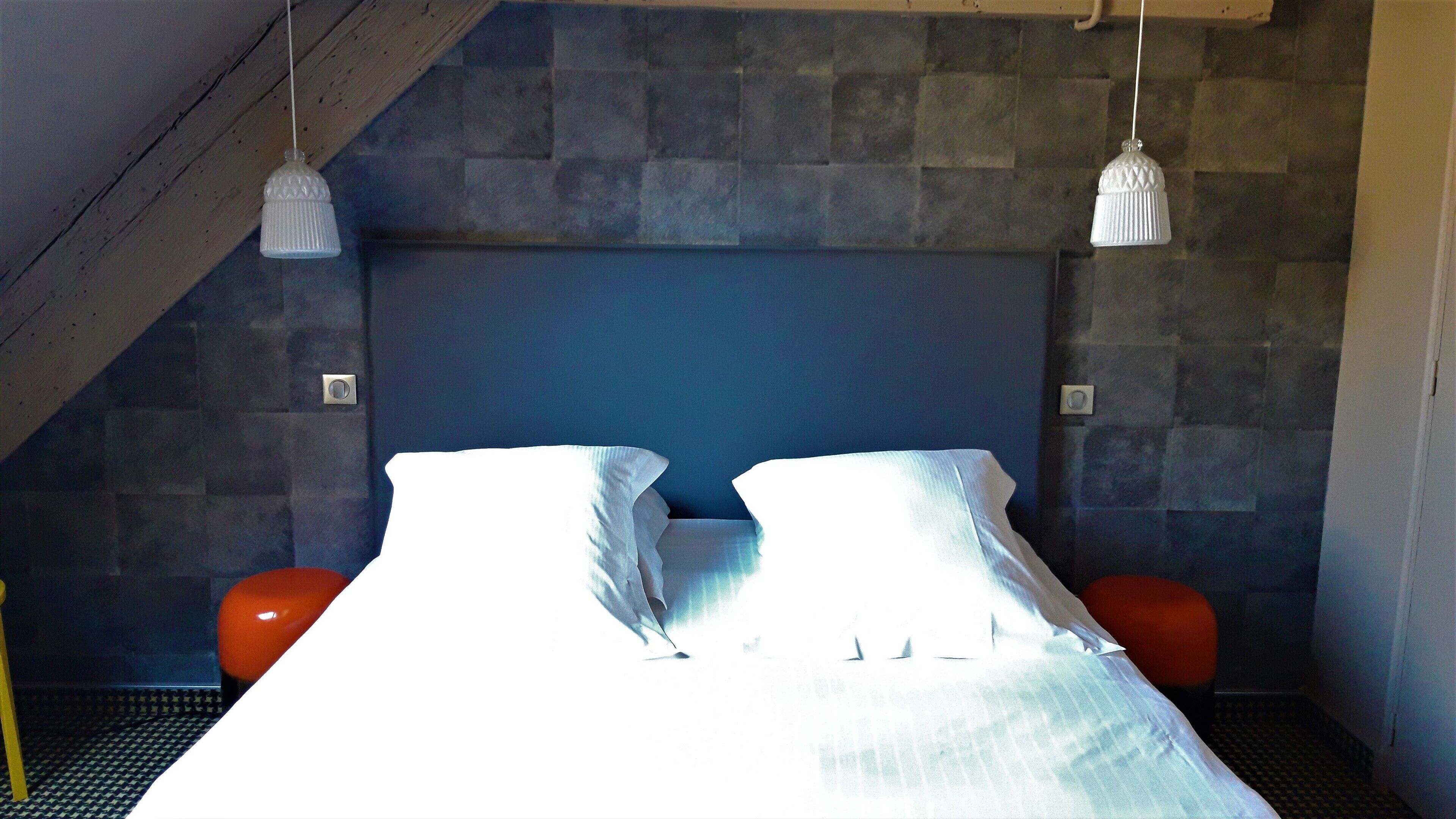 A bedroom with a sloped ceiling, a bed with white linens, two white pendant lights, and a stone wall.