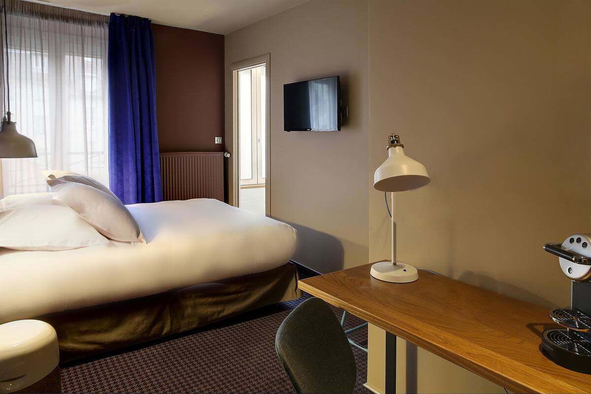 A hotel room with a bed, a desk, a chair, a TV, and a lamp.