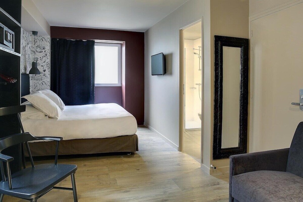 A bedroom with a bed, a chair, a TV, and a door leading to a bathroom.