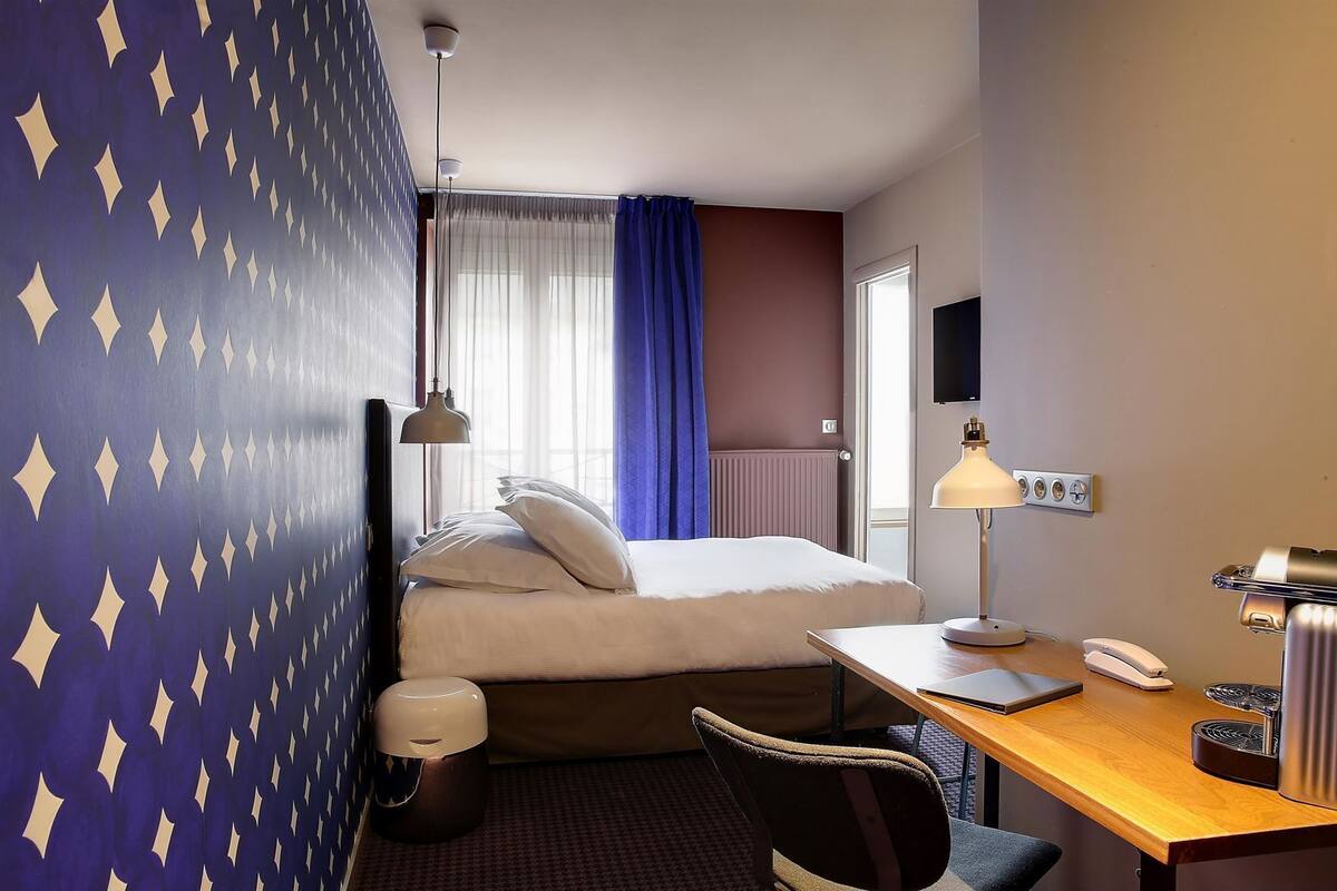 A hotel room with a bed, a desk, a chair, a lamp, a trash bin, and a television.