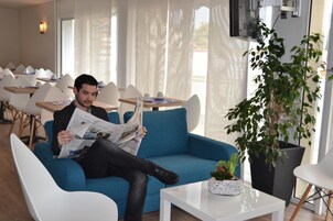 Un hombre leyendo un periódico en un área de descanso moderna con sofás azules, sillas blancas y una planta grande en maceta.
