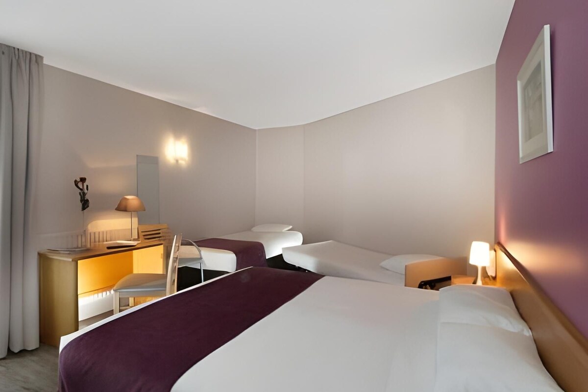 A hotel room with two beds, a desk with a lamp, a chair, and a bedside table with a lamp.