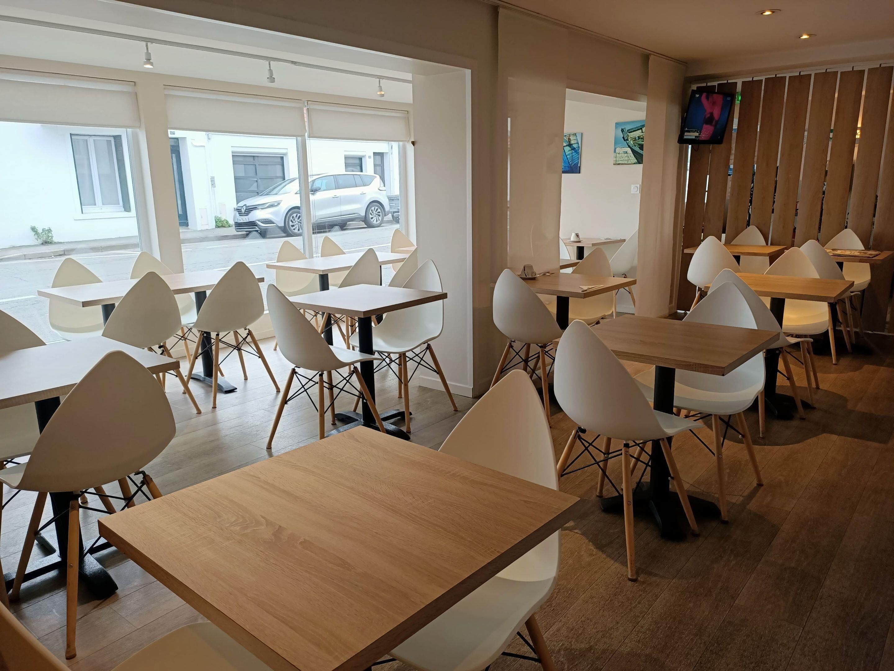 Un comedor moderno con mesas y sillas, ventanas amplias con vista a coches estacionados y un televisor montado en la pared.