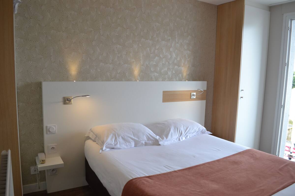 A hotel room with a large bed, a bedside table, a wall-mounted light, and a window with curtains.