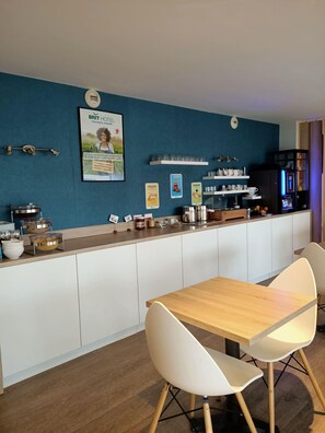 A modern kitchen with a blue accent wall, a coffee machine, and a dining area with a wooden table and white chairs. - Brit Hotel Les Alizés (Pornic)