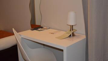 Ein Hotelzimmer mit einem weißen Schreibtisch, einem weißen Stuhl, einer Lampe und einem Bett mit einem orangefarbenen Kissen.