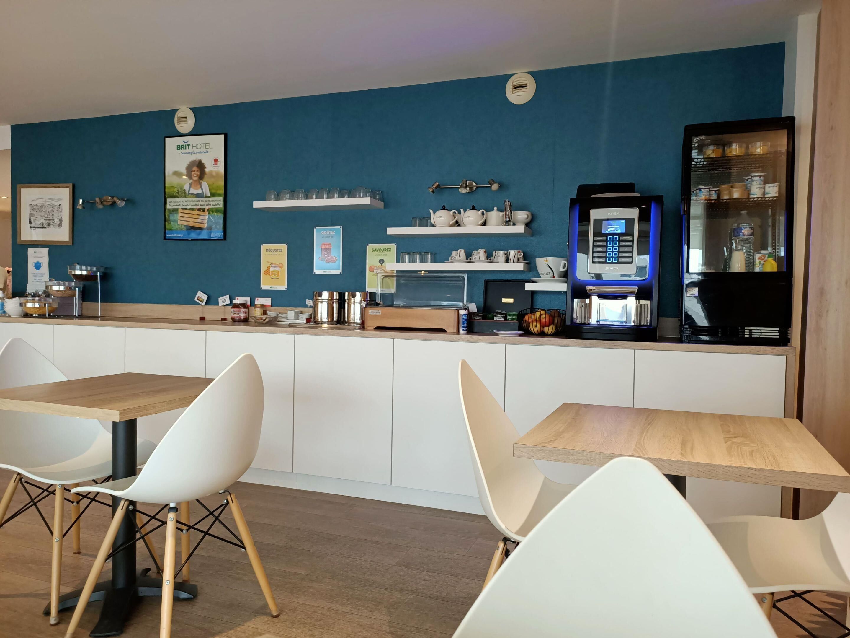 Ein Hotelrestaurant mit weißen Stühlen, einer Theke mit Kaffeeautomaten und einer blauen Wand mit Postern.