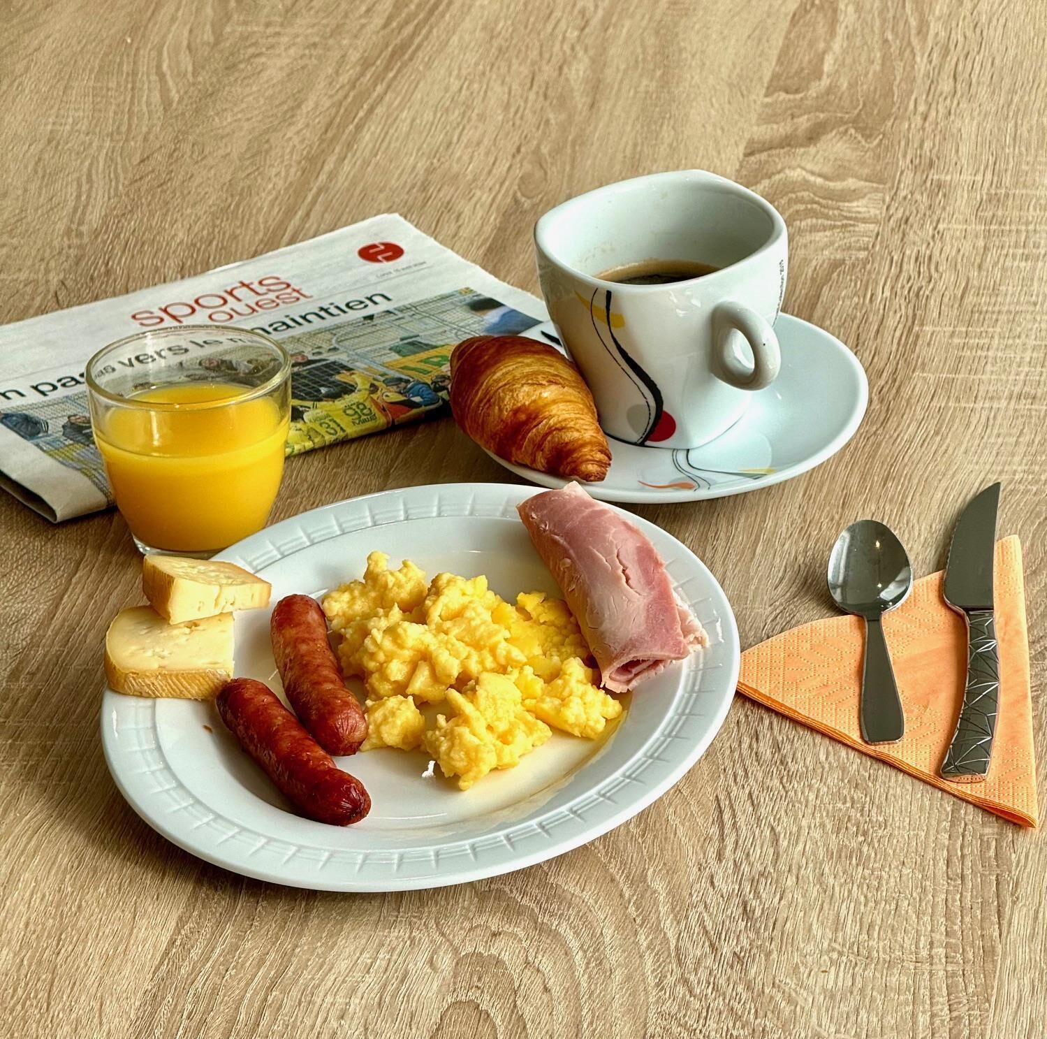 A breakfast plate with scrambled eggs, ham, sausages, cheese, and a croissant, accompanied by a glass of orange juice and a cup of coffee, with a newspaper on the table.