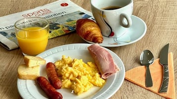 A breakfast plate with scrambled eggs, ham, sausages, cheese, and a croissant, accompanied by a glass of orange juice and a cup of coffee, with a newspaper on the table.