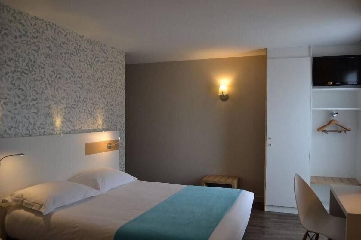 A hotel room with a bed, a desk, a chair, a TV, and a wall-mounted light.