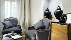 A modern hotel room with a large window, two armchairs, a wooden side table, and a minibar. - Mario de Fiori 37 (Rome)