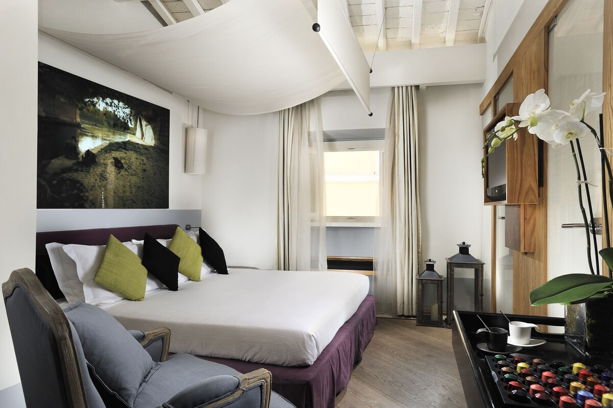 A hotel room with a bed, a chair, a table with a cup, and a painting on the wall.