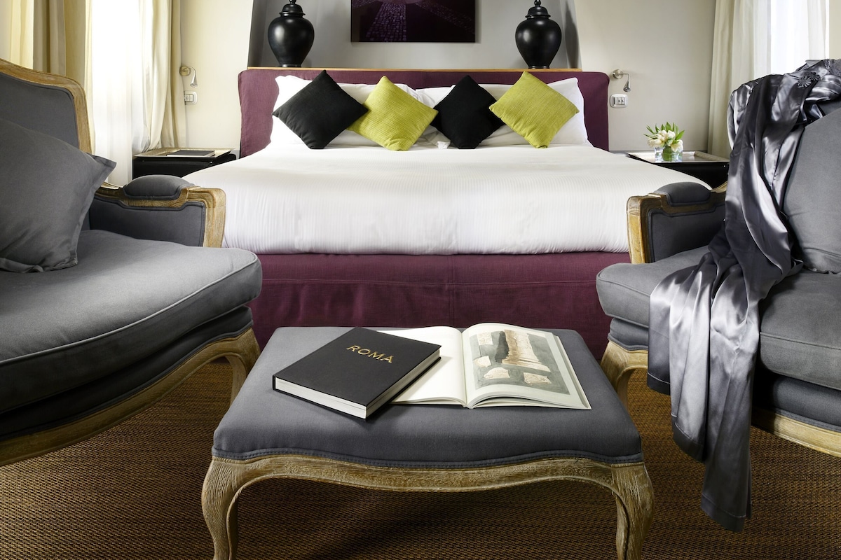 A hotel room with a large bed, two armchairs, a small table with a book, and a painting on the wall.