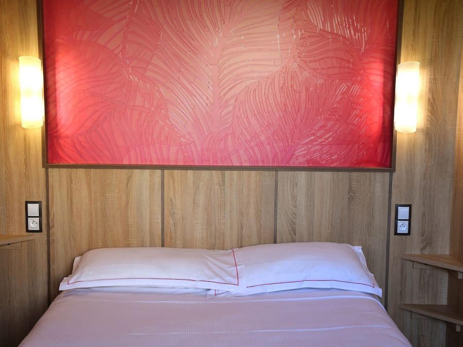 A neatly made bed with white linens and red piping, a large red abstract painting on the wall, and wooden paneling.