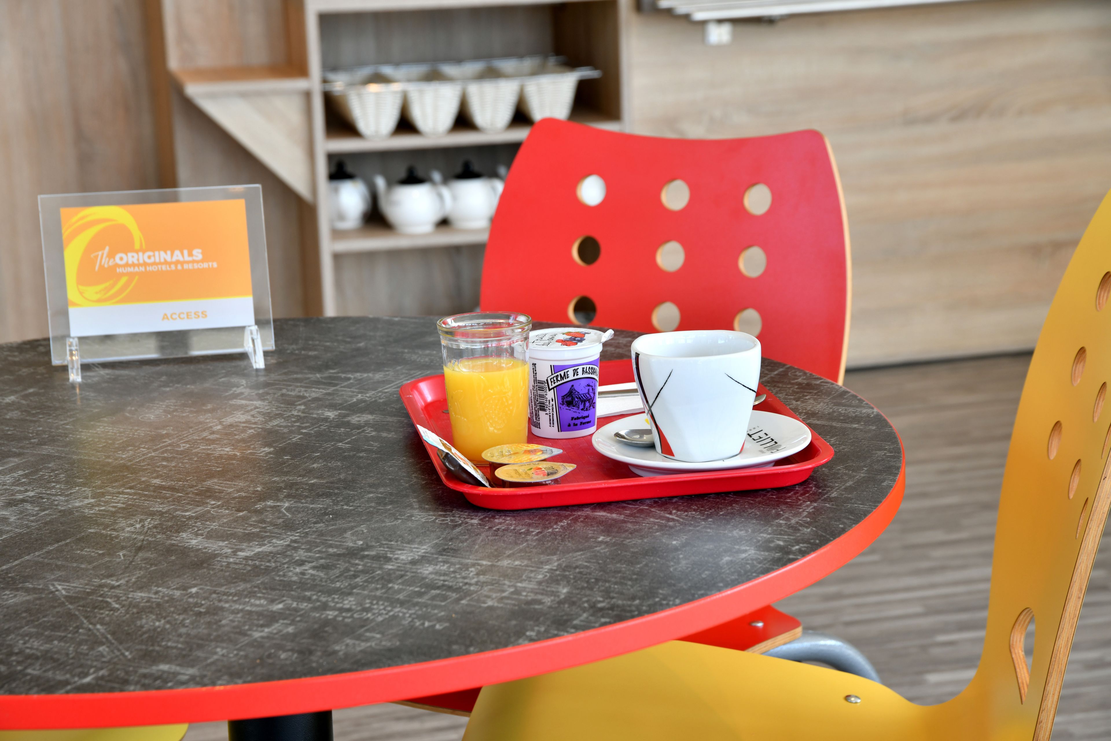 Een ontbijfwagen met jus d'orange, een yoghurtbeker en een witte kop op een ronde tafel met een rode stoel.
