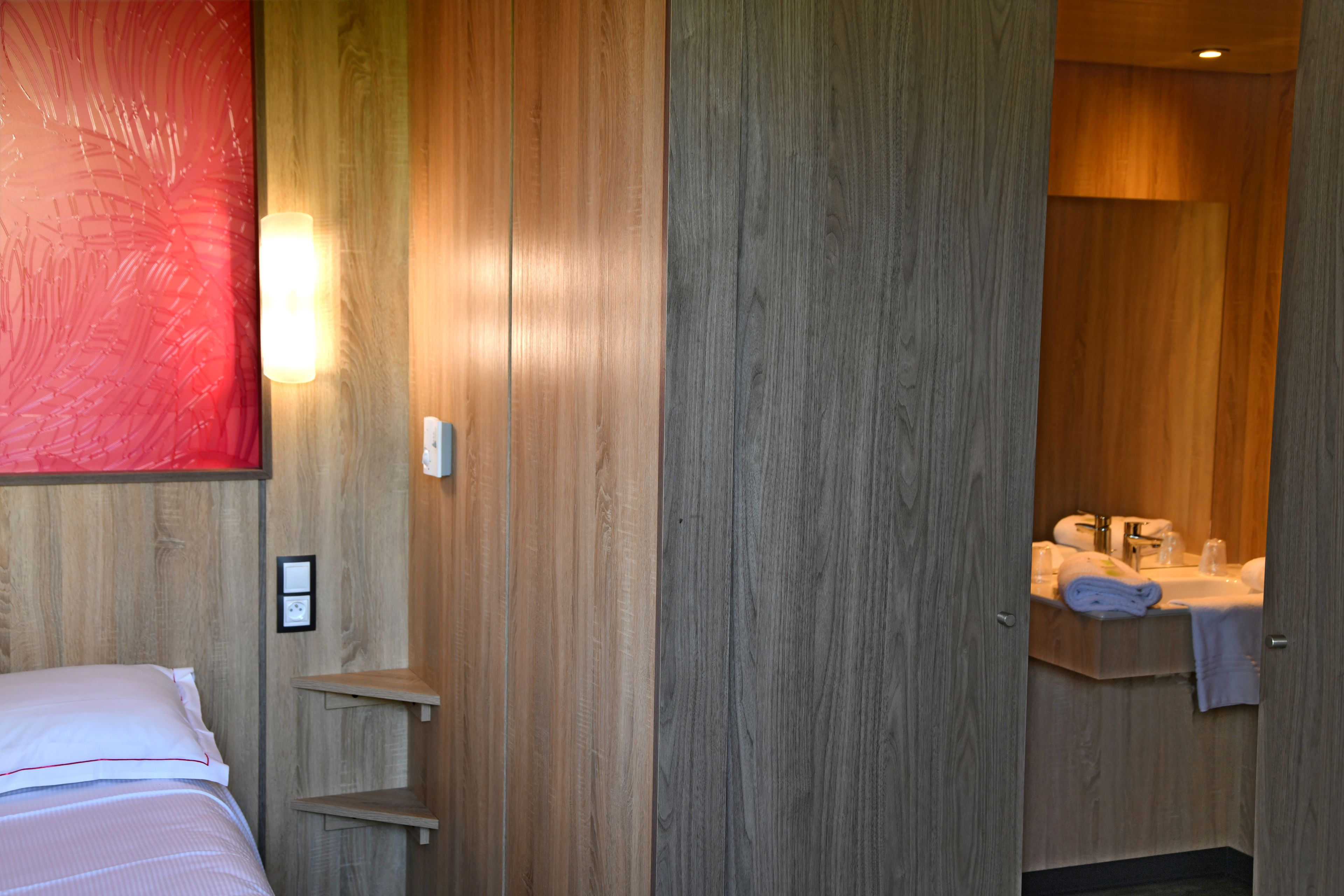 Habitación de hotel con pared de madera, cama con ropa blanca, mesita de noche pequeña y baño visible a través de una puerta abierta.
