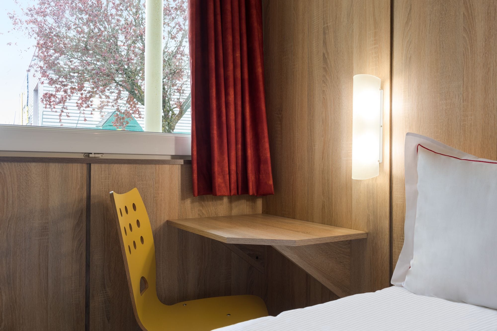 Habitación de hotel con pared de madera, cortina roja, silla amarilla, mesita de noche y almohada blanca.