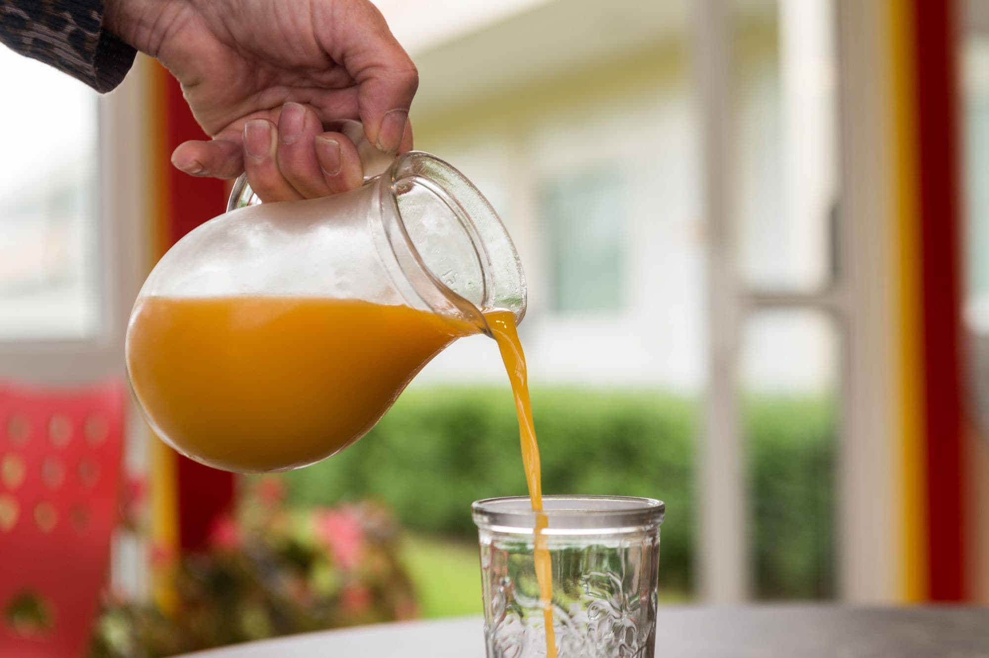A hand pouring orange juice from a glass pitcher into a clear glass.