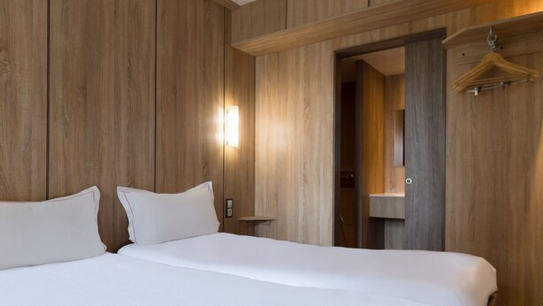 Habitación de hotel con pared de madera, cama con ropa blanca, mesita de noche y perchero.