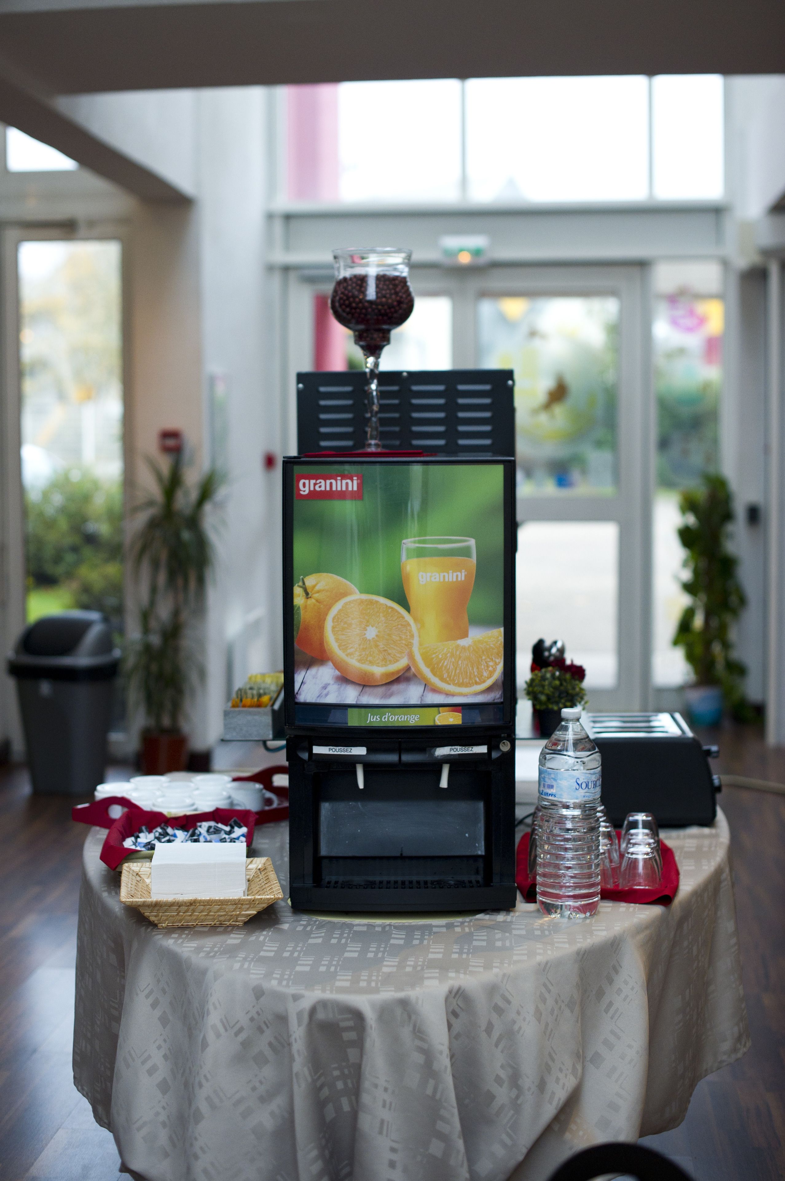 A self-service beverage dispenser with a granini advertisement, a bottle of water, and a basket of pastries on a table in a well-lit room with large windows.