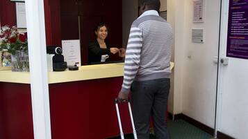Zona de recepción de hotel con mostrador, recepcionista y un huésped con maleta.