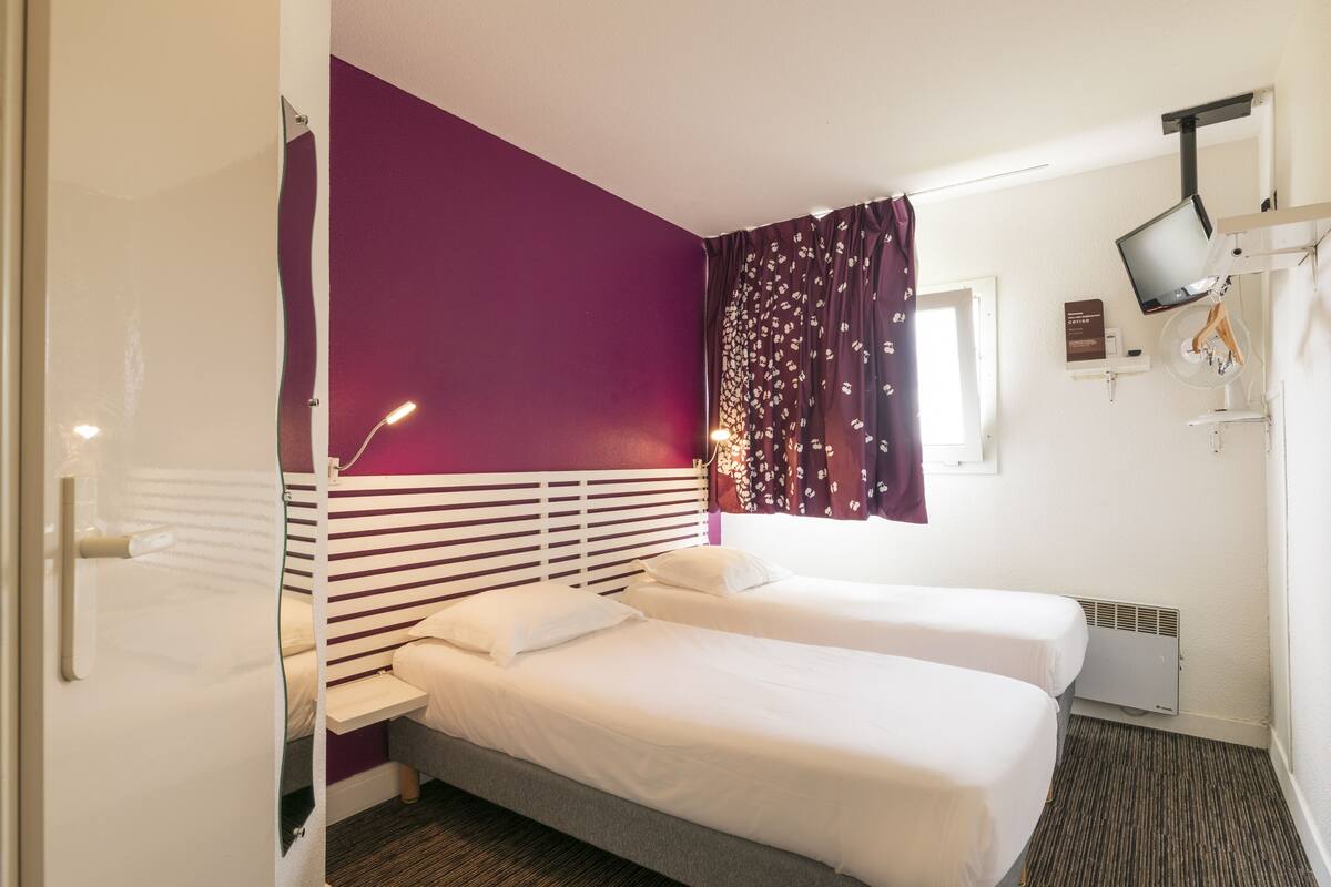 A hotel room with two beds, a TV, a window with curtains, and a bedside table.