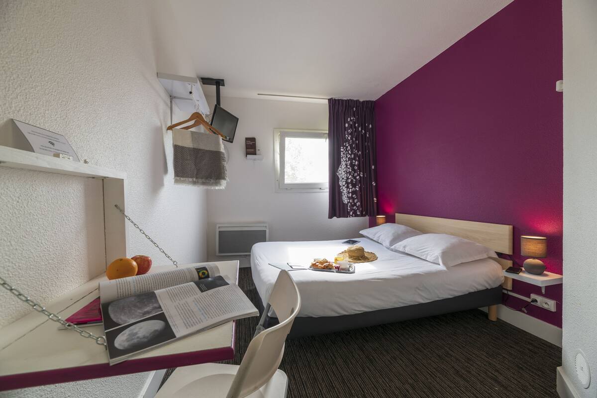A hotel room with a bed, a desk with a chair, a small table with a lamp, and a window with curtains.