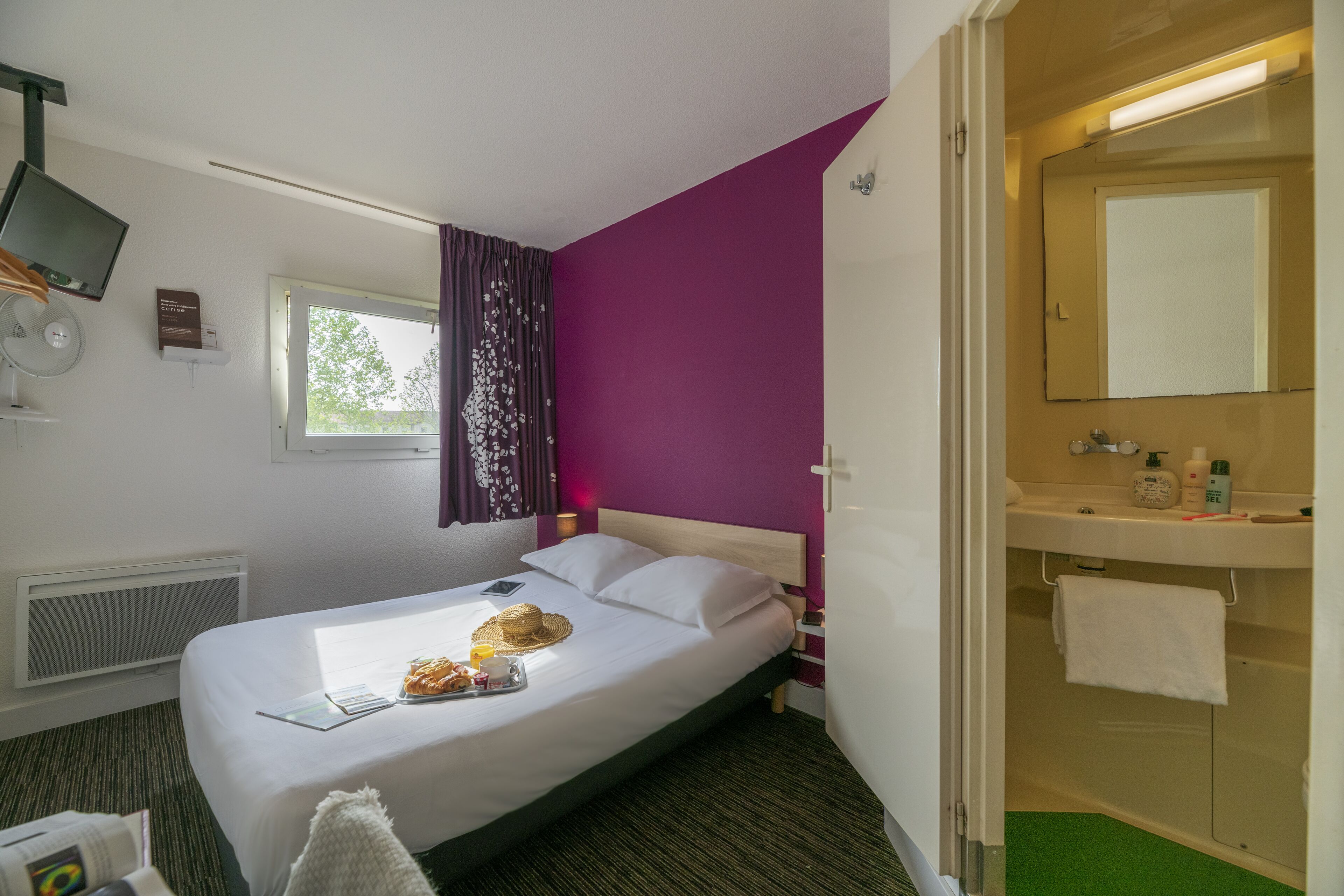 A hotel room with a bed, a window with curtains, a bedside table with a lamp, a mirror, and a bathroom with a sink and towel rack.