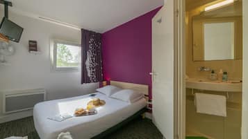 A hotel room with a bed, a window with curtains, a bedside table with a lamp, a mirror, and a bathroom with a sink and towel rack.