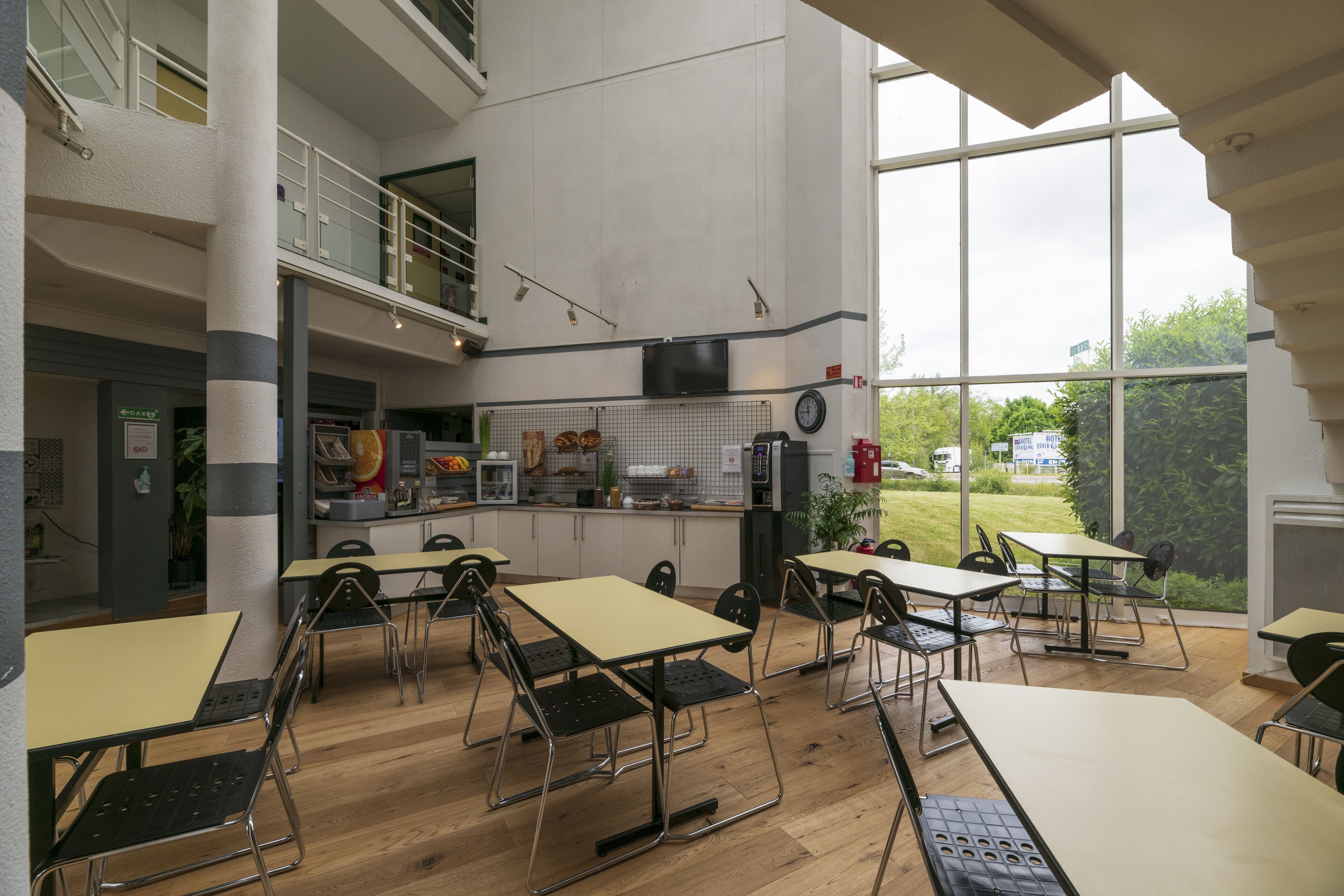 A modern indoor dining area with multiple tables and chairs, a kitchen area with appliances, and large windows offering a view of the outdoors.