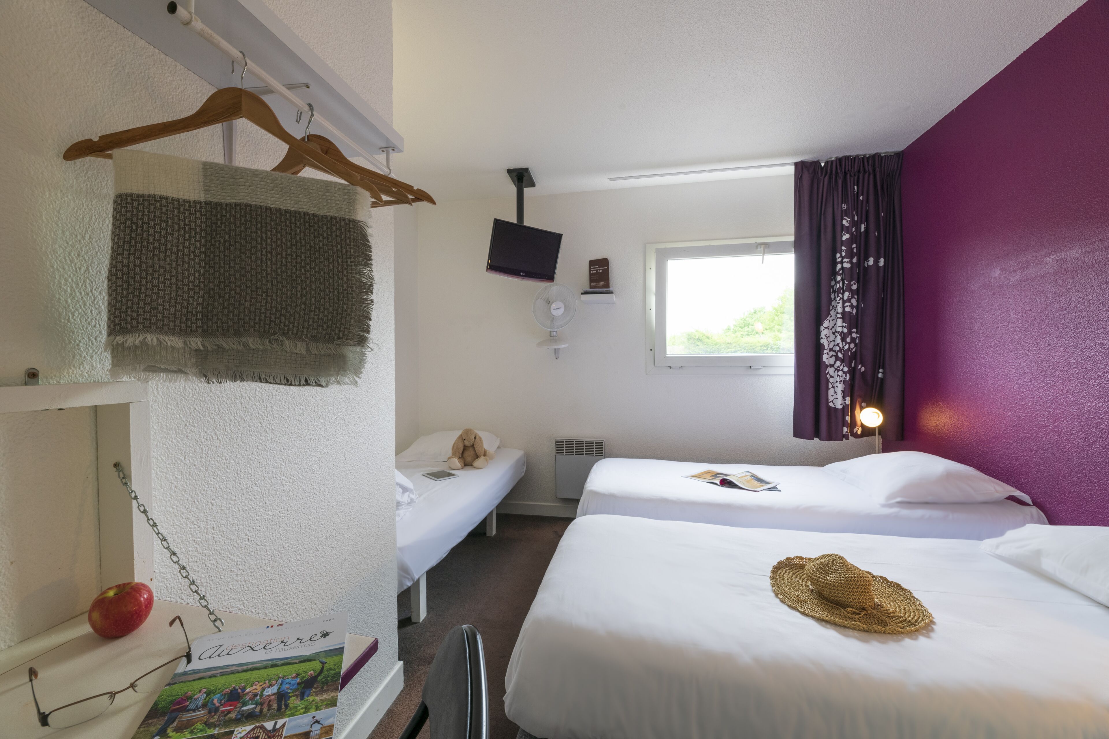 A hotel room with two beds, a TV, a window with curtains, and a small desk with a book and glasses.