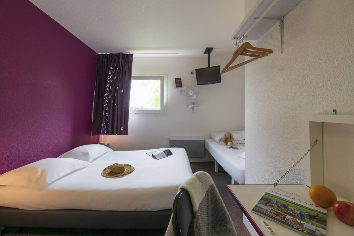 A hotel room with a bed, a bedside table with a book and fruit, a small table with a chair, a TV mounted on the wall, and a window with curtains.