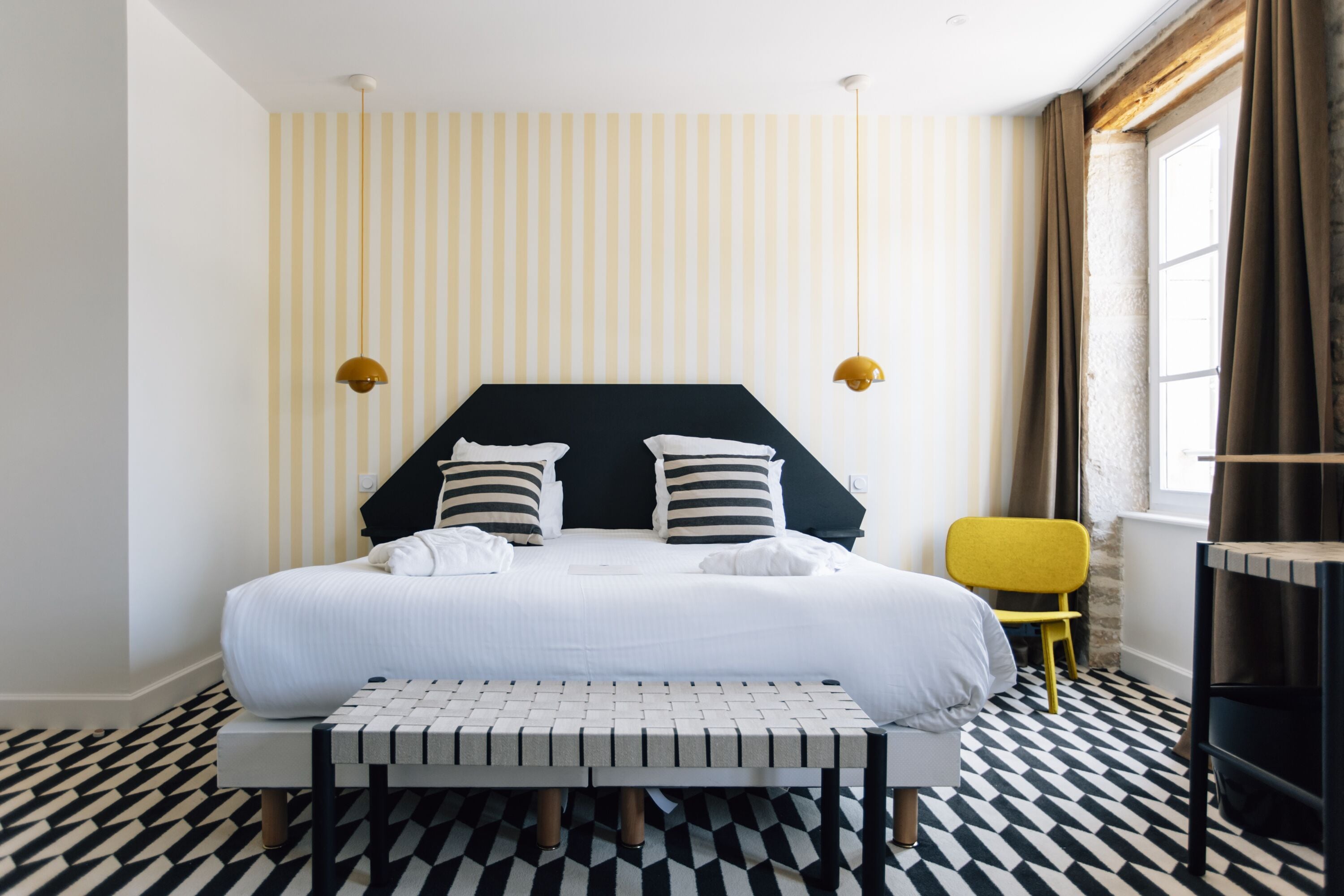 A hotel room with a bed, a bench, a chair, and a window with curtains.