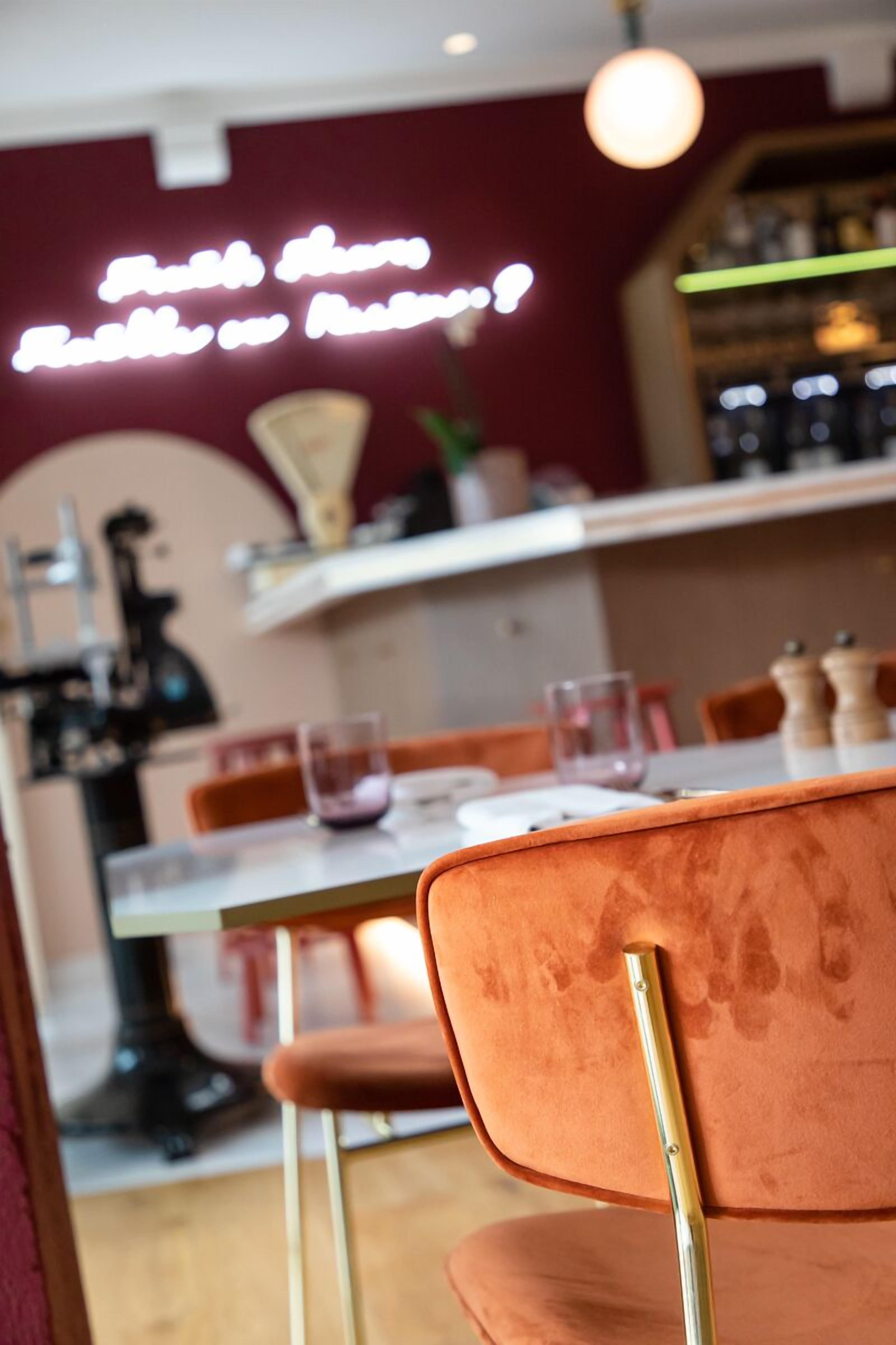 A restaurant interior with a red wall, a neon sign, a table with chairs, and a bar area in the background.