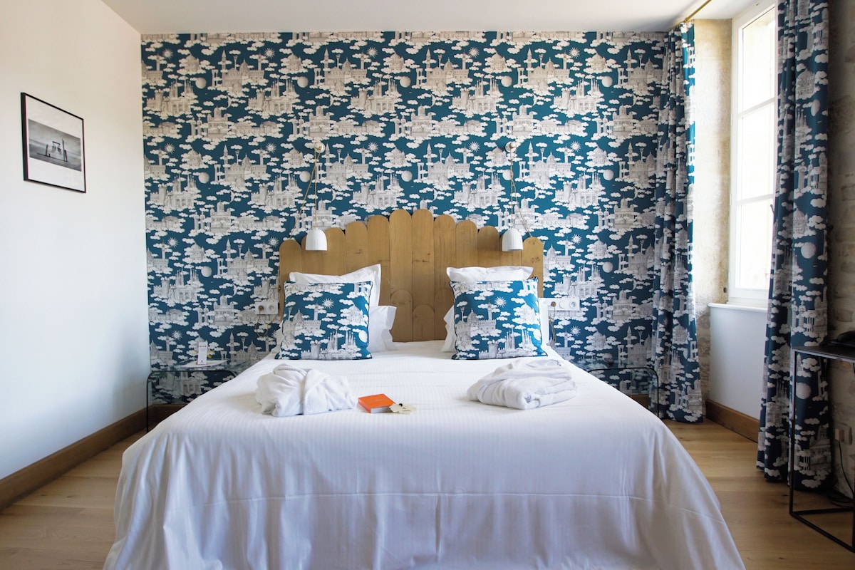 Una habitación de hotel con una cama grande, ropa de cama blanca, un cabecero de madera y un papel tapiz estampado.