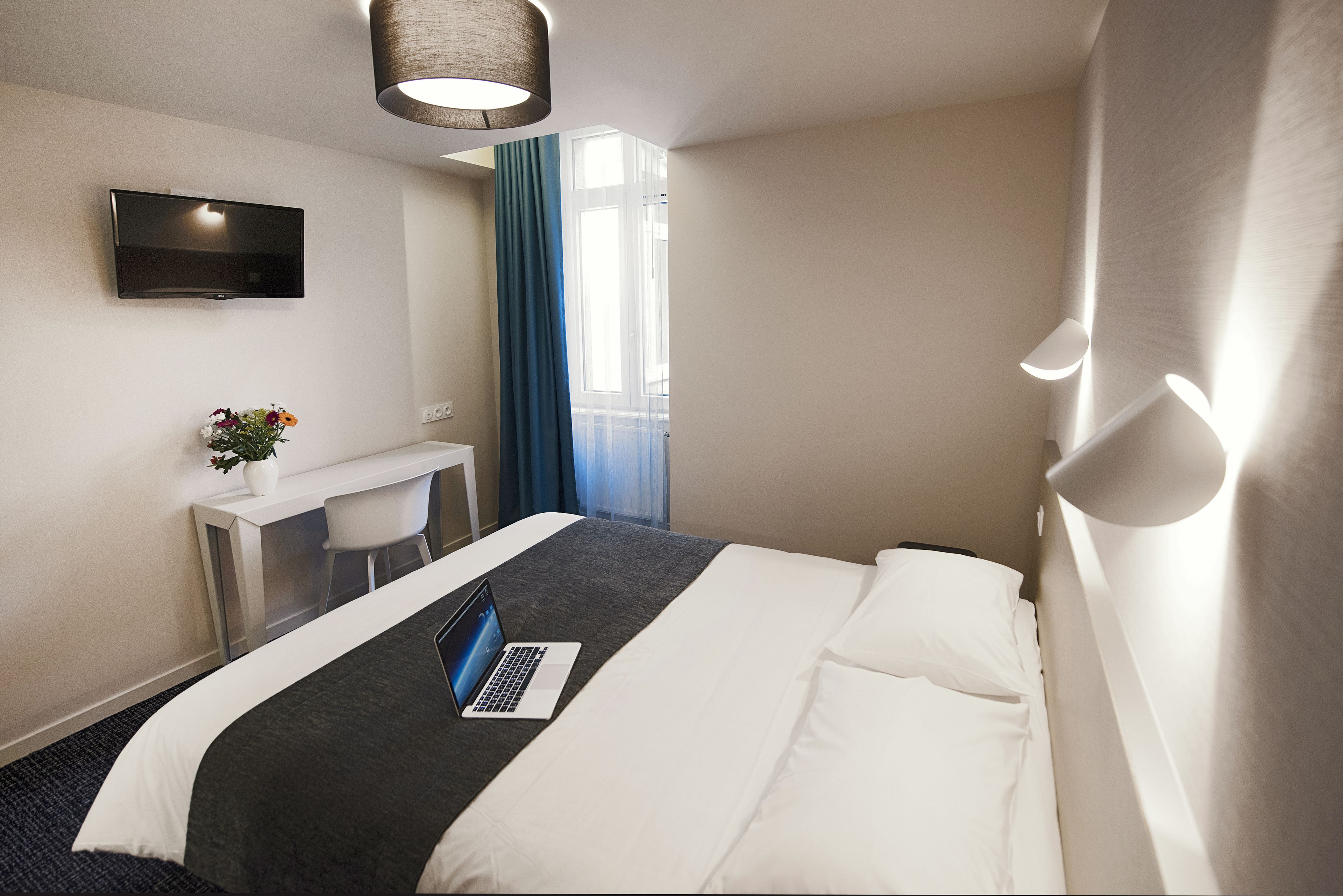 A hotel room with a bed, a desk with a laptop, a TV, and a window with curtains.