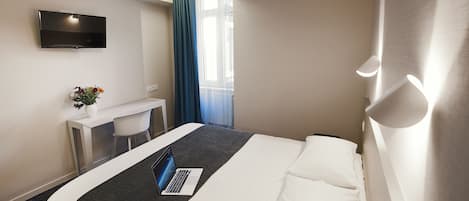 A hotel room with a bed, a desk with a laptop, a TV, and a window with curtains.
