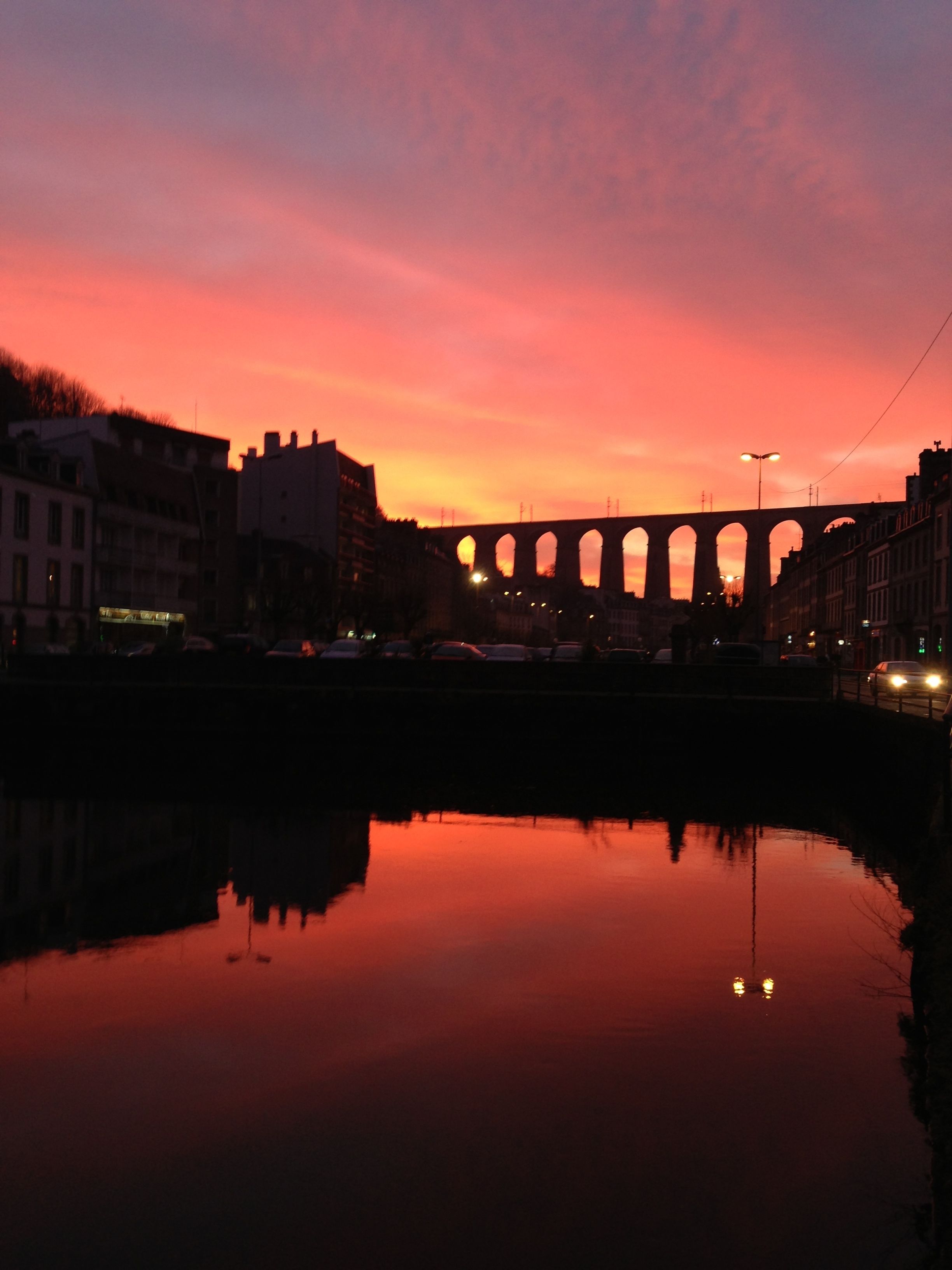 Ein Sonnenuntergang über einer Brücke, flankiert von Gebäuden, mit einem ruhigen Fluss, der den Himmel spiegelt.